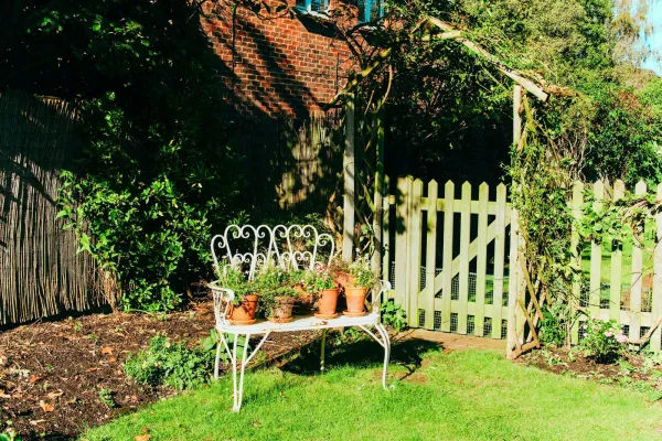 Weiße Gartenbank mit Terrakottatöpfen vor einem Gartentor im Sonnenlicht